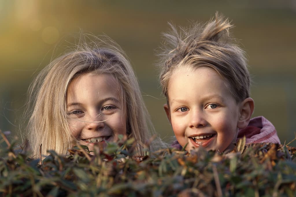 zwei Kinder schauen lachend hinter einem Busch hervor