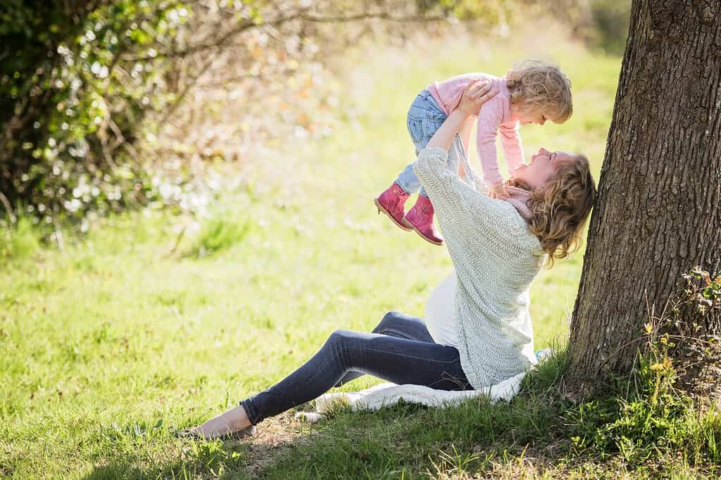 eine schwangere Frau sitzt unter einem Baum und hält lachend ihr Kind in die Höhe.