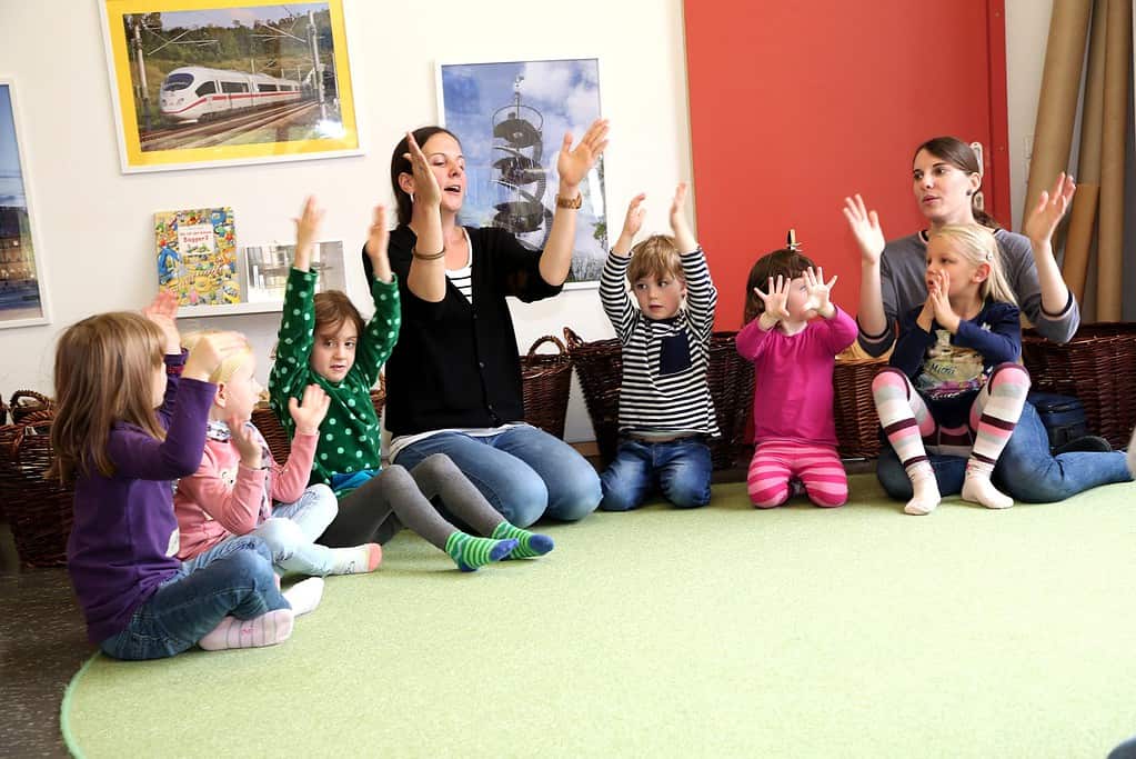 Kinder und Erzieherinnen sitzen im Kreis auf dem Boden und Singen mit Handbewegungen