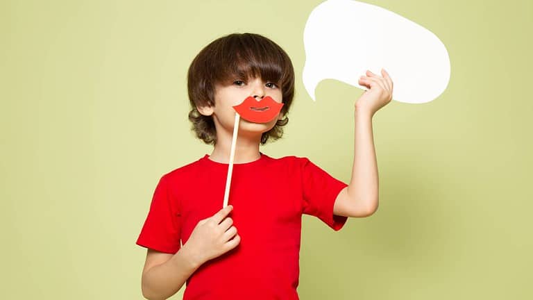 ein Kind hält in einer Hand ein Schild mit roten Lippen vor dessen Mund und in der anderen Hand hält es eine Sprechblase hoch