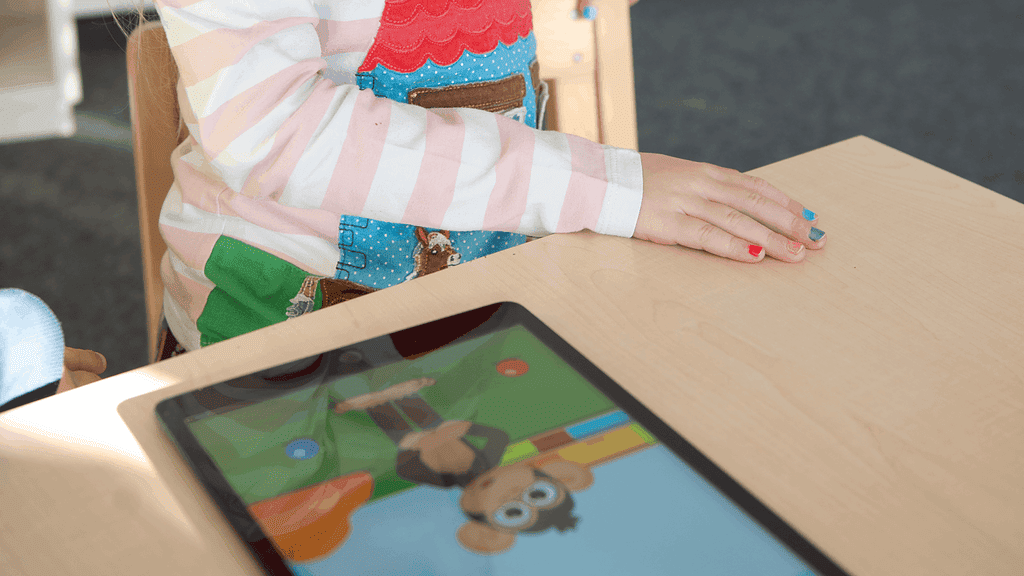 ein Kind sitzt am Tisch, auf dem ein Tablet liegt, welches die Kinderbefragung abspielt
