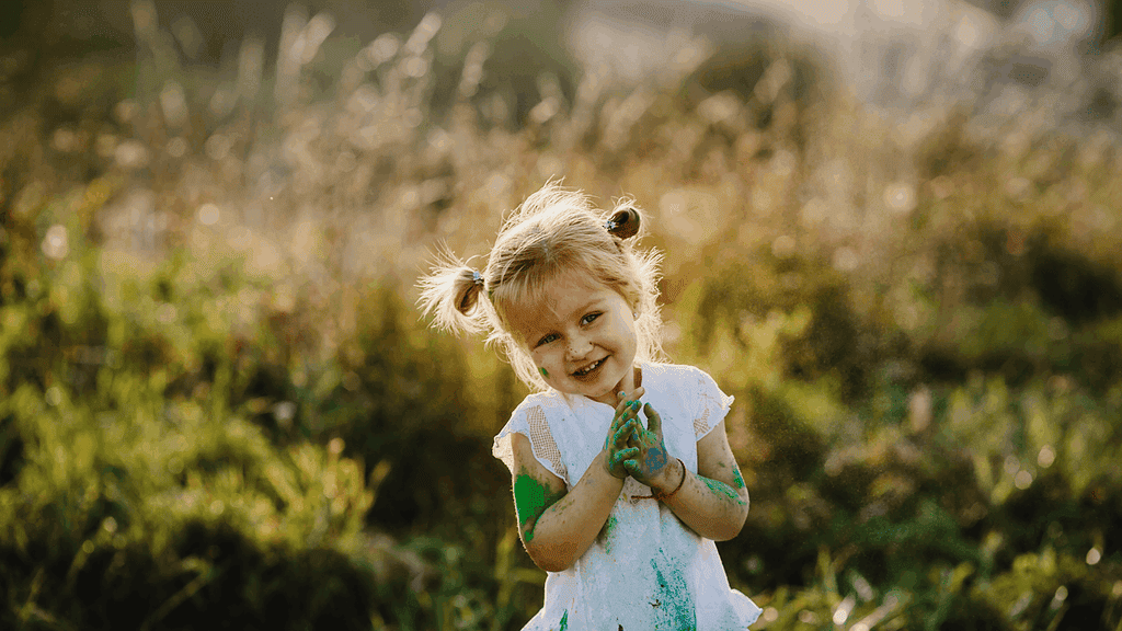 ein Kind steht lächelnd in der Natur mit bunten Händen vor dem Körper zusammen gehalten